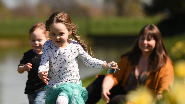 Two young children run past daffodils with a lady out of focus sat behind them.